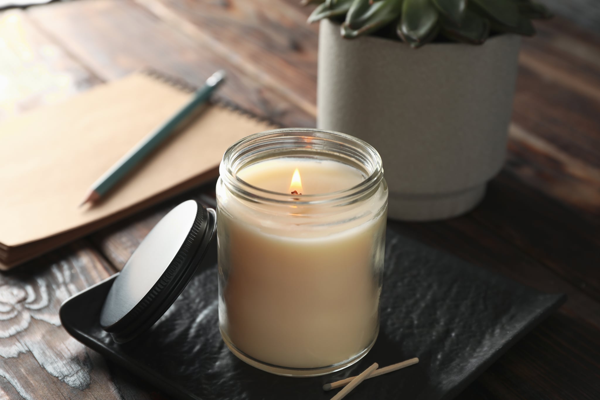 Burning candle in glass jar, succulent and notebook on wooden background, close up