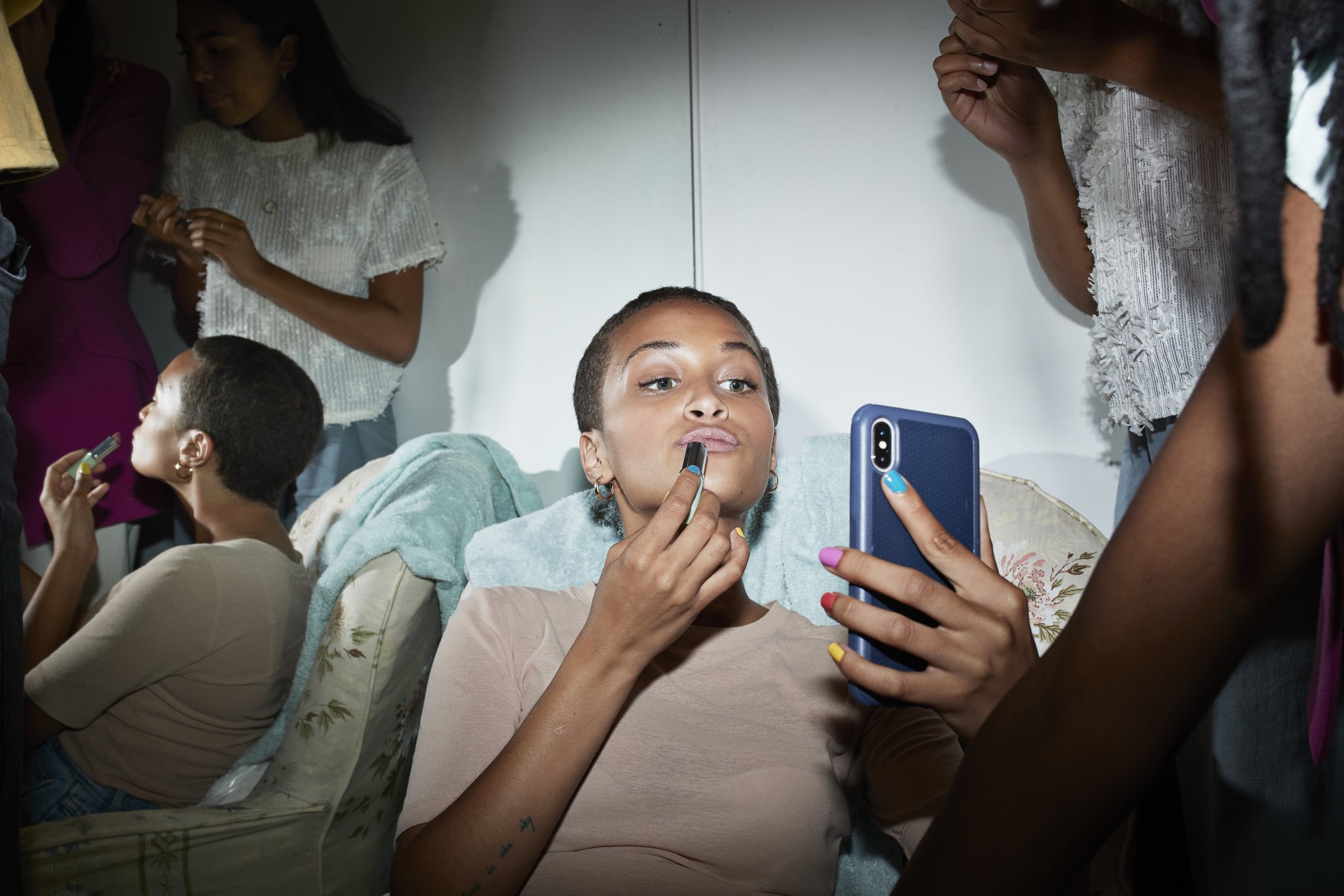 Woman applying lipstick while looking in mobile phone with friends reflecting in mirror at home