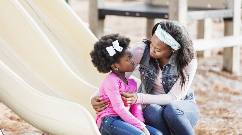 A young African-American woman helping her 5 year old daughter who is sitting on a slide on a playground with a sad expression on her face.