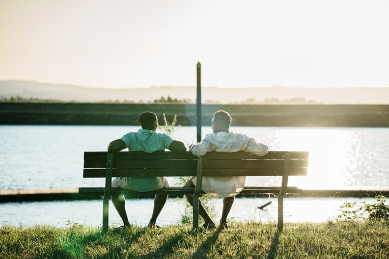 Two black men sit on a park bench and cheerfully chat while watching the sun set over a river after spending the afternoon fishing together.