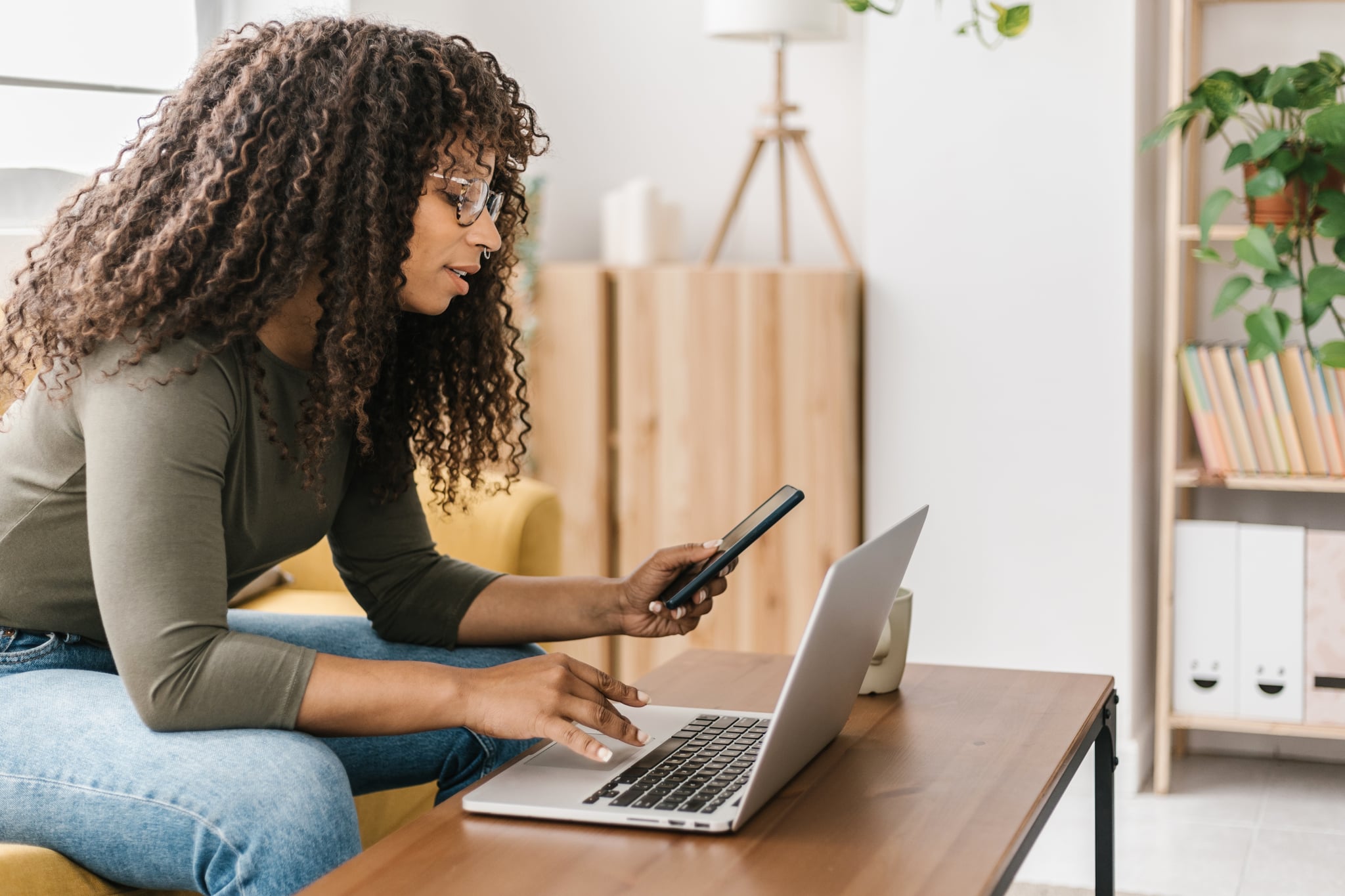Young Black woman using smartphone while typing on laptop at home