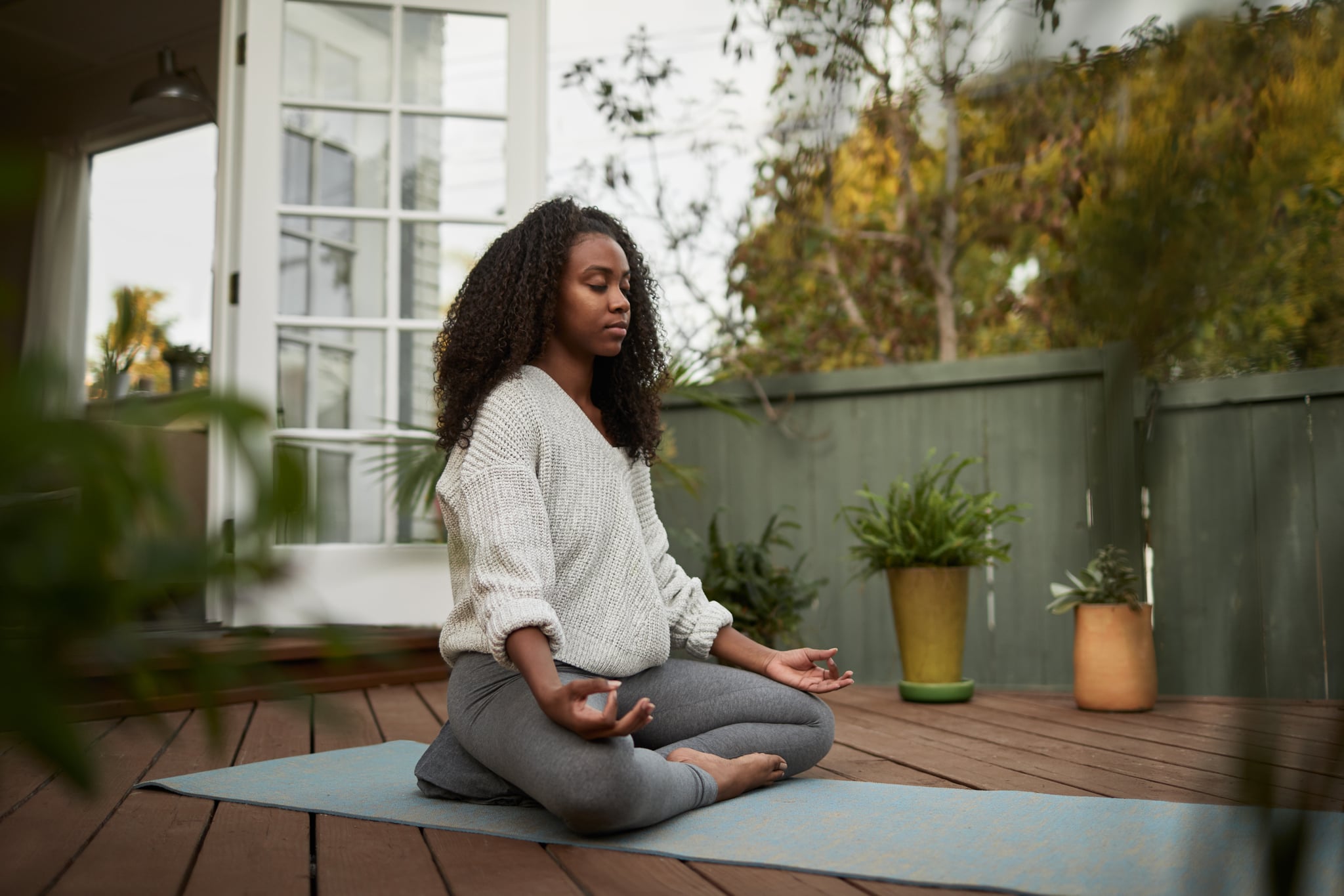 Young African American woman sitting on exercise mat outside on her patio and meditating in the lotus pose during a yoga session