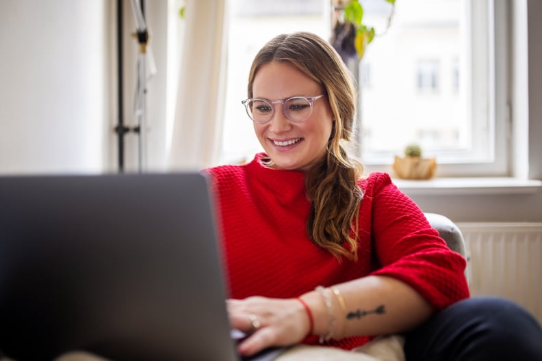 幸福的女人坐在沙发上,致力于笔记本电脑。微笑的女性在家工作。