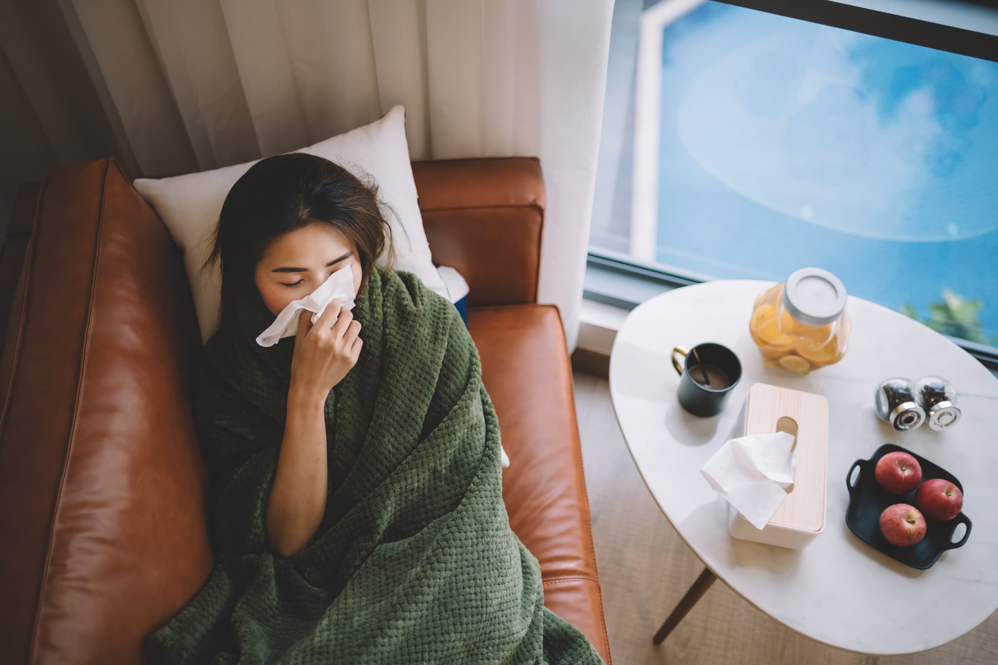 an sick asian chinese female lying on sofa in living room covered with blanket and wiping nose with tissue paper