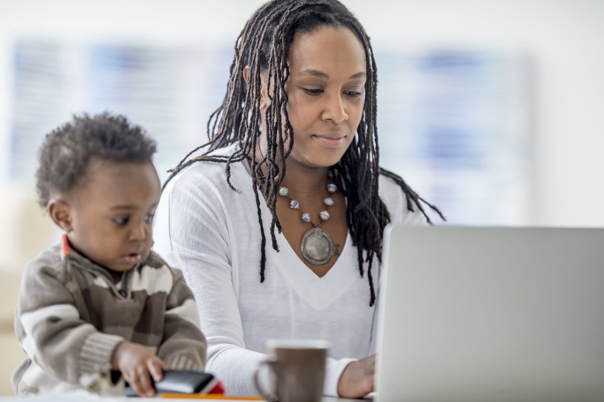 A woman of African descent is at home with her baby boy. She is working from home on her laptop. Her son is playing with a phone beside her.