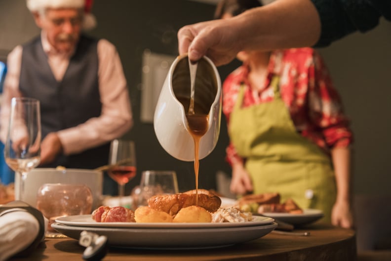 Gravy being poured from a jug onto a plate of Christmas lunch. A woman is wearing an apron out of focus in the background and there are more plates and glasses on the table ready for the family to sit down.