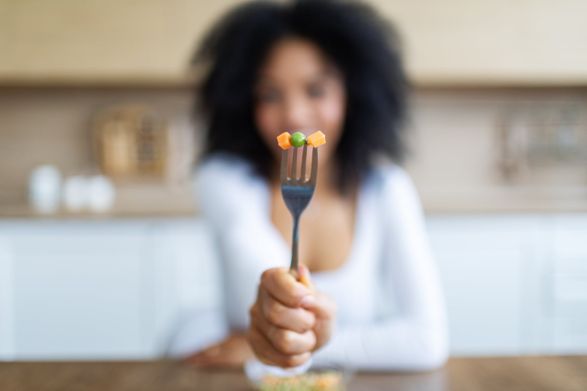 Young woman eating vegetables, showing fork with green peas and carrot.