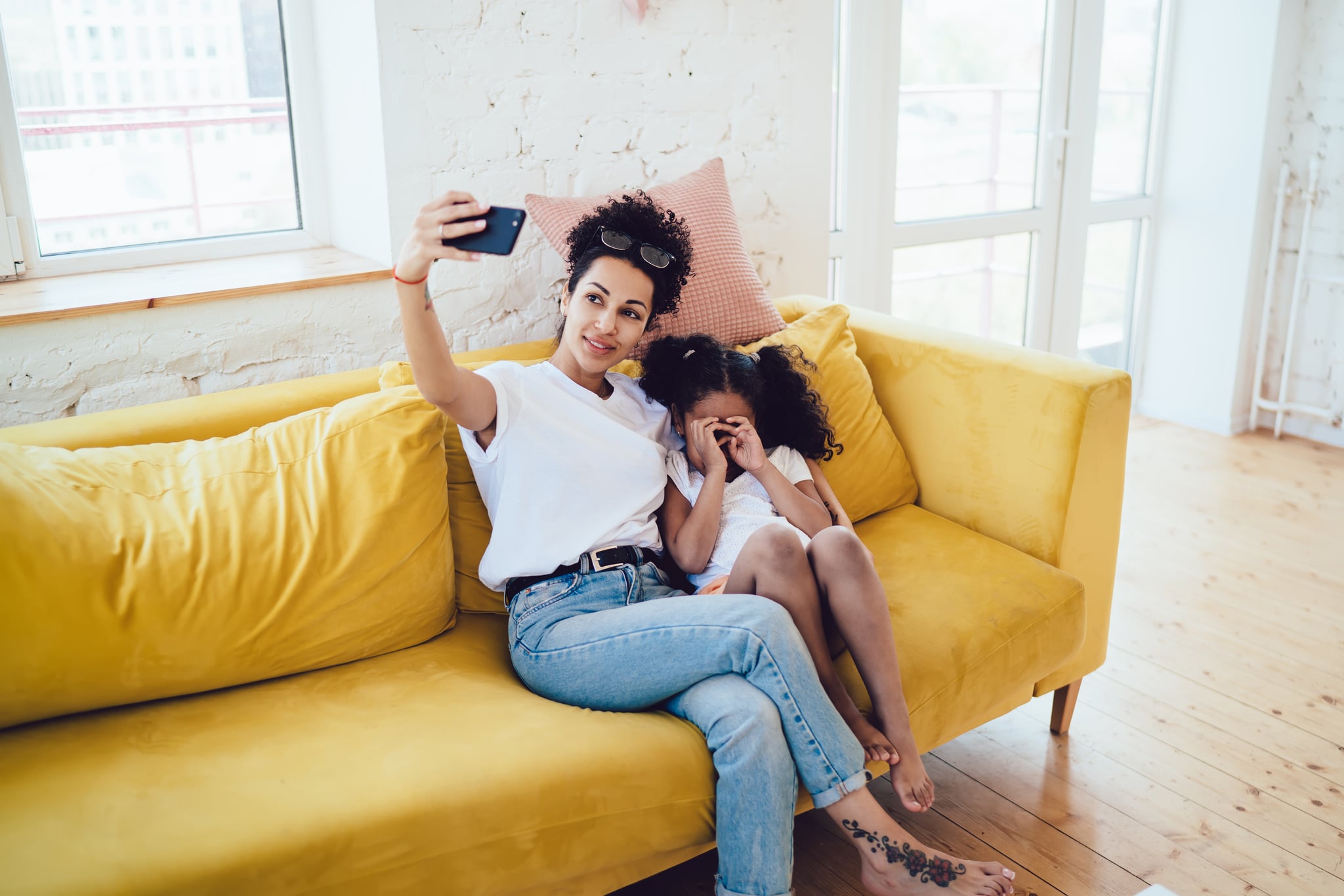 Cheerful smiling African American young woman taking selfie with curly adorable daughter hiding face from camera with hands on yellow cosy couch at home