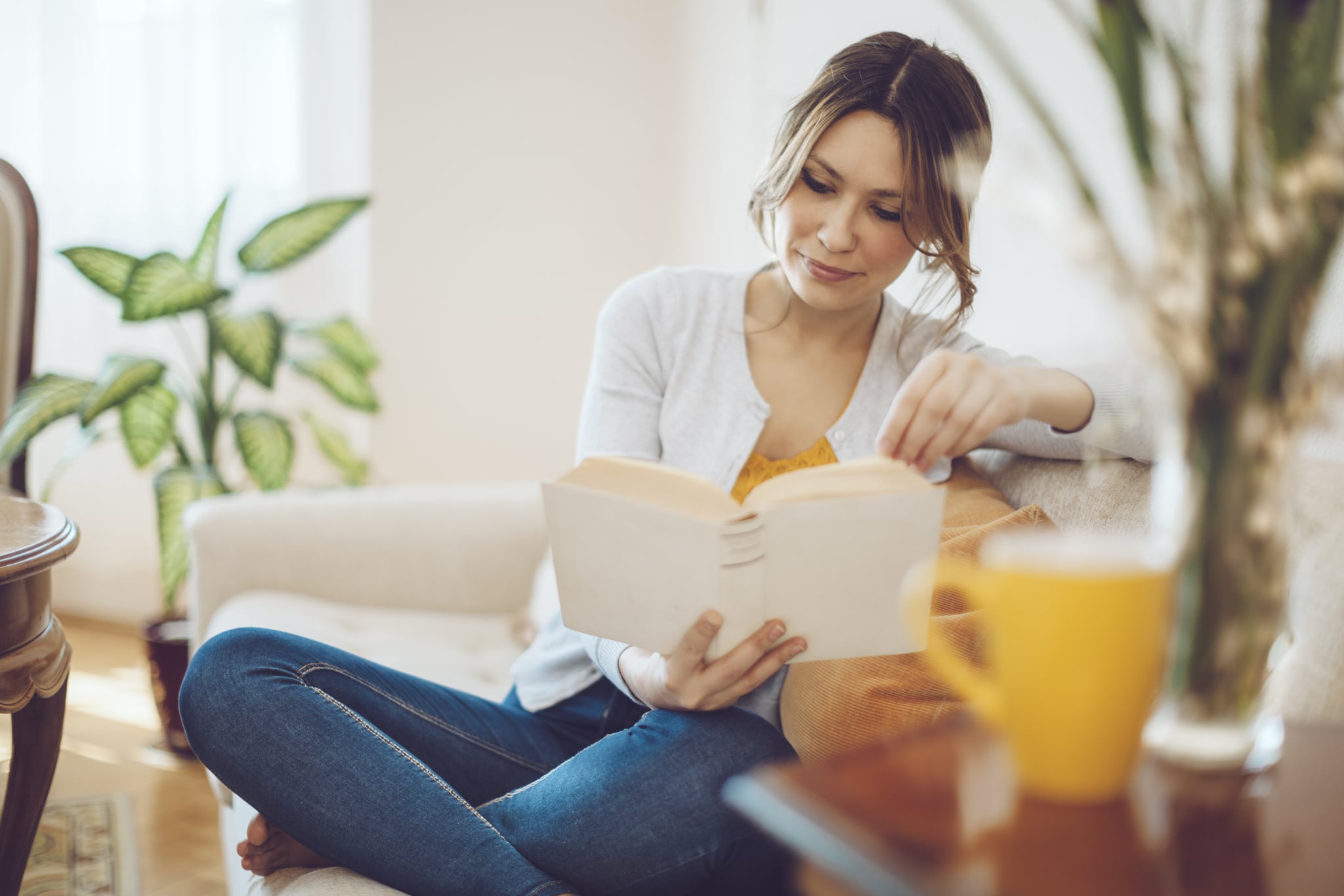 Content woman is reading a book on a white sofa at home. She is sitting comfortable and is wearing blue jeans and a grey sweater. The living room is cosy and elegant. Her hair and eyes are brown. Candid shot.