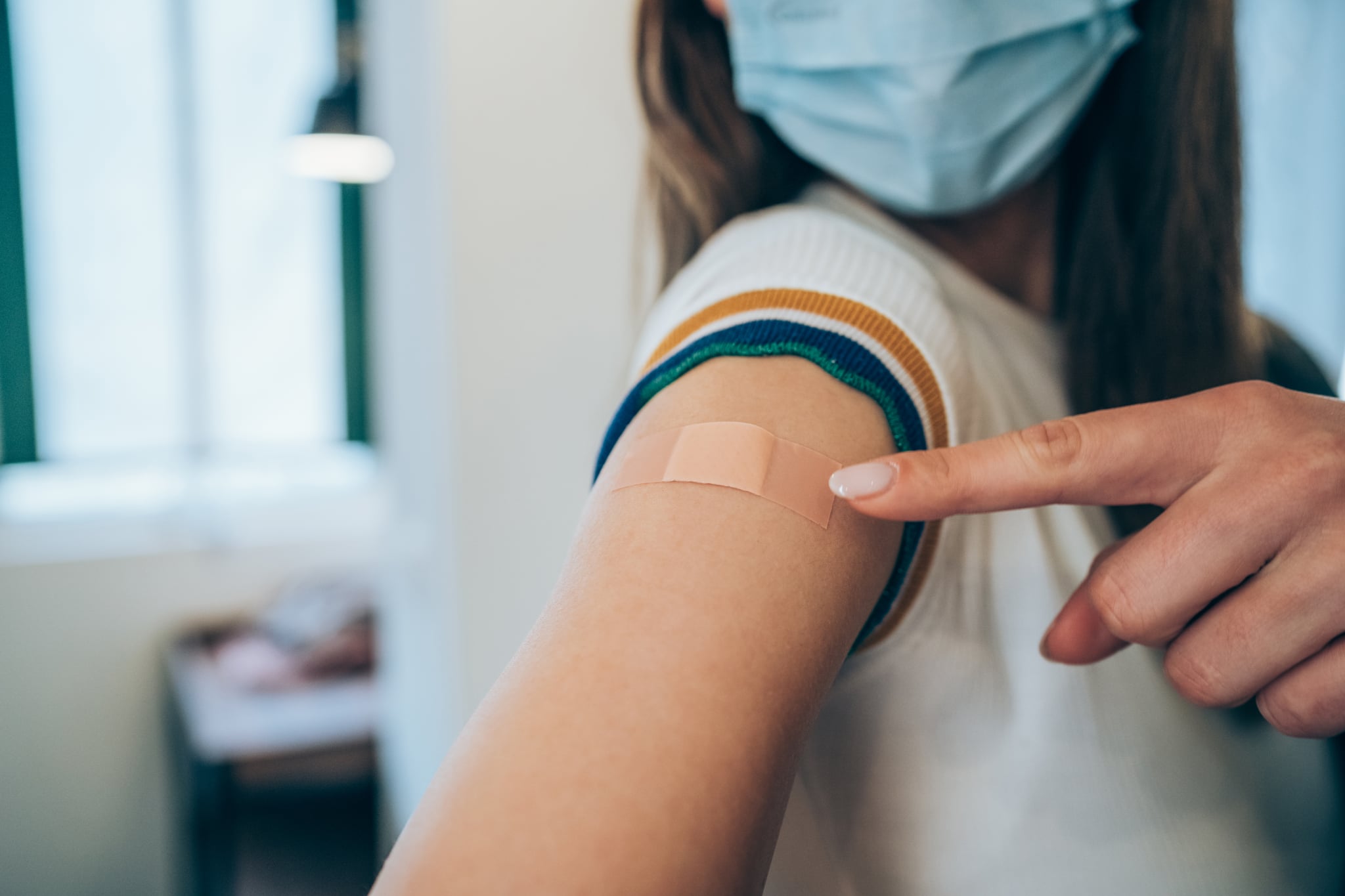 Woman wearing protective face mask and pointing at her arm with a bandage after receiving COVID-19 vaccine. Young woman showing her shoulder after getting coronavirus vaccine at vaccination center.