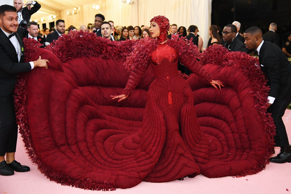 OK, everyone at the 2019 Met Gala needs to move over right now. No, seriously, Cardi B took the theme "Camp: Notes on Fashion" (aka, be as extra as possible) to the extreme, and her dress takes up the entire pink carpet. She went from rapper to vintage peacock goddess in a custom, wine-red Thom Browne gown with a 10-foot train that rippled out behind her and had to be carried by multiple full-grown men. Someone sure knows how to make an entrance!
Cardi's gown was hand-embroidered with 30,000 dyed coque feathers and a full-coverage headpiece codesigned by Browne and Stephen Jones. The sequin heart cut-out on the back of the dress and the carefully placed ruby nipples on the front of the gown make the entire look feel like a sexy red shrug made for the goddess of love herself. 
"I designed this dress for Cardi specifically because she has the ultimate beauty in a woman's body, and that is what the dress is about for me: taking advantage of that beauty," Browne told Vogue. Not only did Cardi's dress accentuate all of her best features, but it definitely put the "red" in red carpet this year. 

    Related:

            
            
                                    
                            

            Mary-Kate and Ashley Olsen Are Peak "Olsen Twins" at the Met Gala, but We&apos;re Not Mad