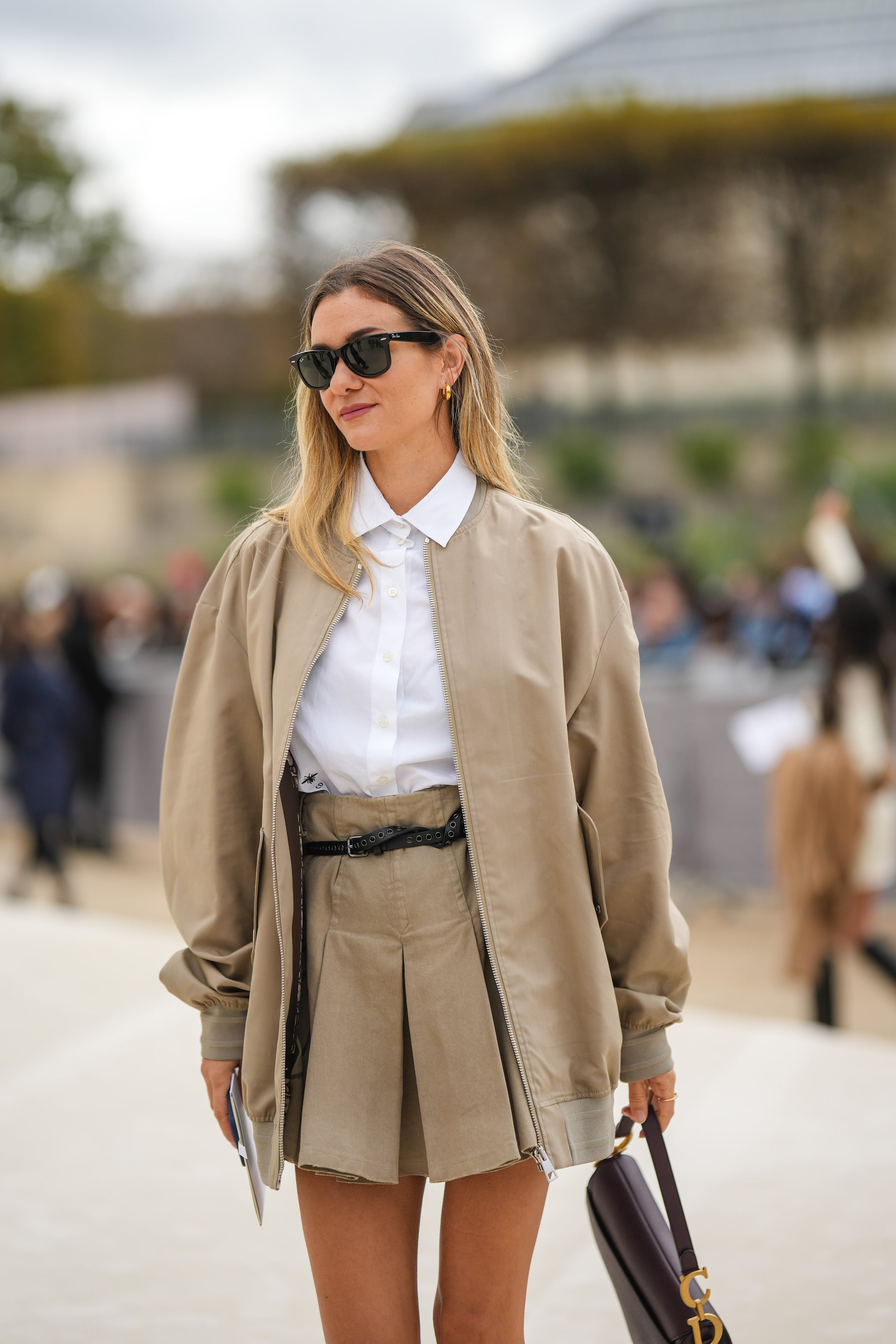 PARIS, FRANCE - SEPTEMBER 27: A guest wears black sunglasses from Ray Ban, gold earrings, a white shirt, a beige jacket from Dior, a matching beige pleated / accordion short skirt from Dior, a black shiny leather belt, a brown shiny leather Saddle handbag from Dior, outside Dior, during Paris Fashion Week - Womenswear Spring/Summer 2023, on September 27, 2022 in Paris, France. (Photo by Edward Berthelot/Getty Images)