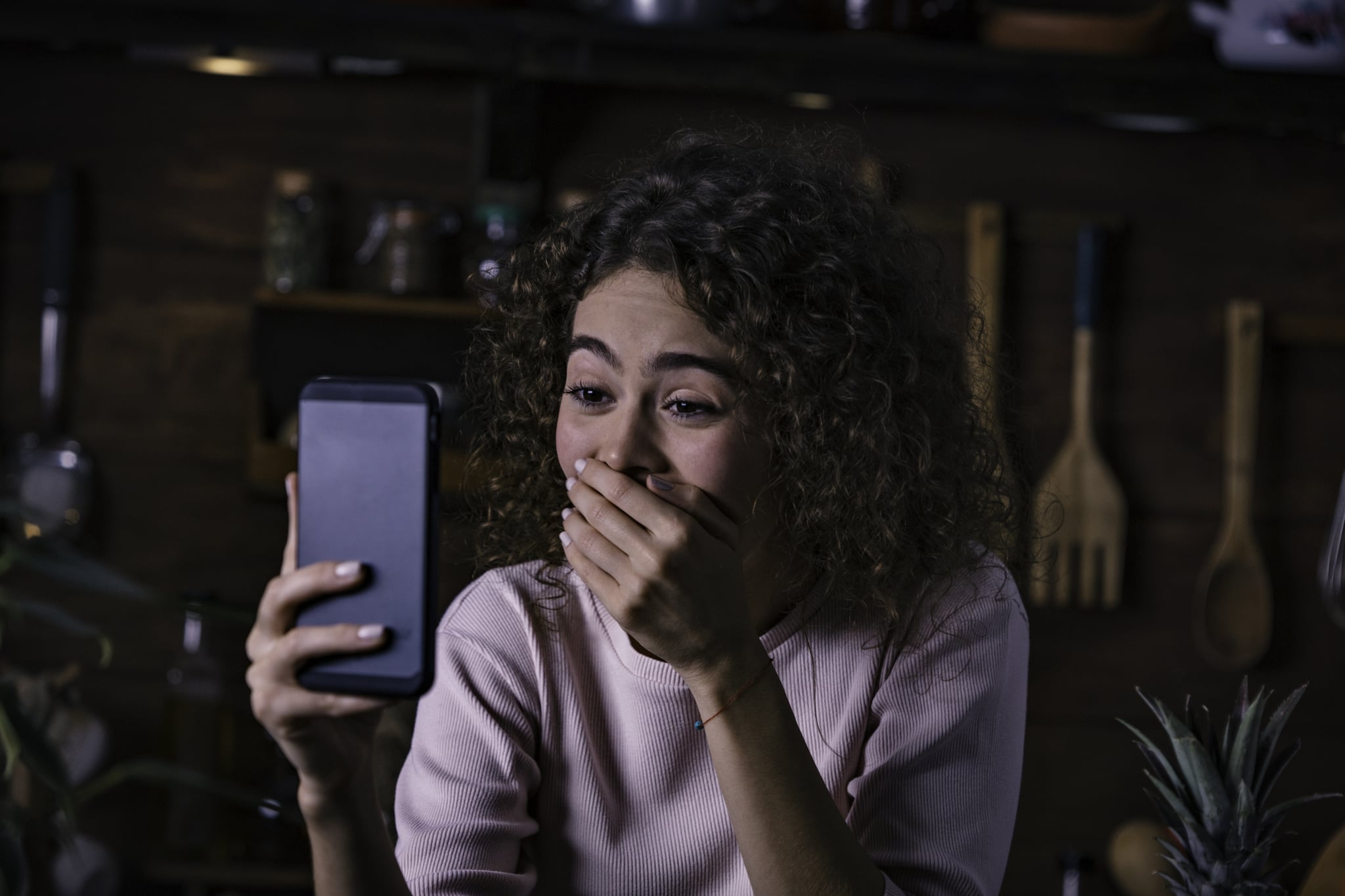 Hispanic curly hair young woman making video call with a friend on a cellphone in a rustic kitchen