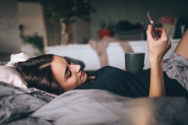 Young woman lying on a bed in her bedroom, using smart phone and drinking first morning coffee
