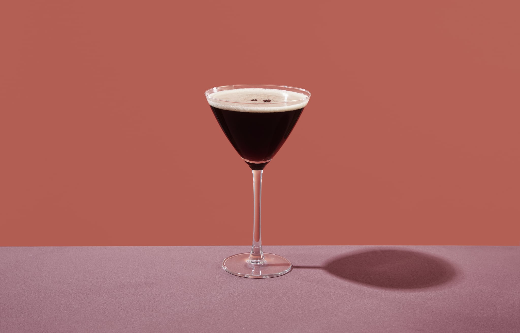 an espresso martini served in a cocktail glass, on a table set with a maroon tablecloth, against a coral pink background